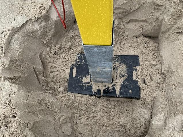 Nahaufnahme eines gelben Pfostens in einem Sandfundament mit nassen Sandkörnern und einer schwarzen Metallplatte.