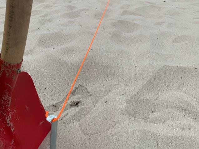 Nahaufnahme eines roten Schaufelblatts neben einem Metallsteg und einer leuchtend orangefarbenen Schnur auf Sand.