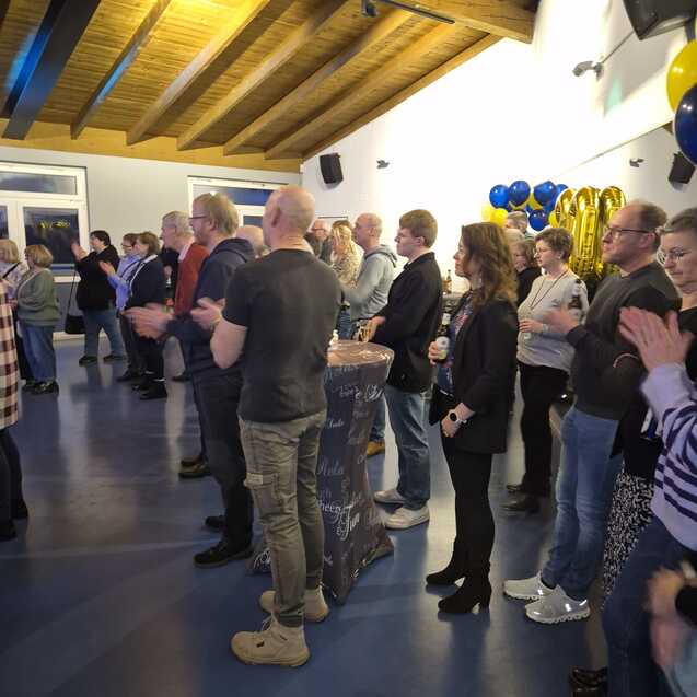 Viele Personen stehen in einem Raum mit blauem Fußboden und hören einem Vortrag zu.