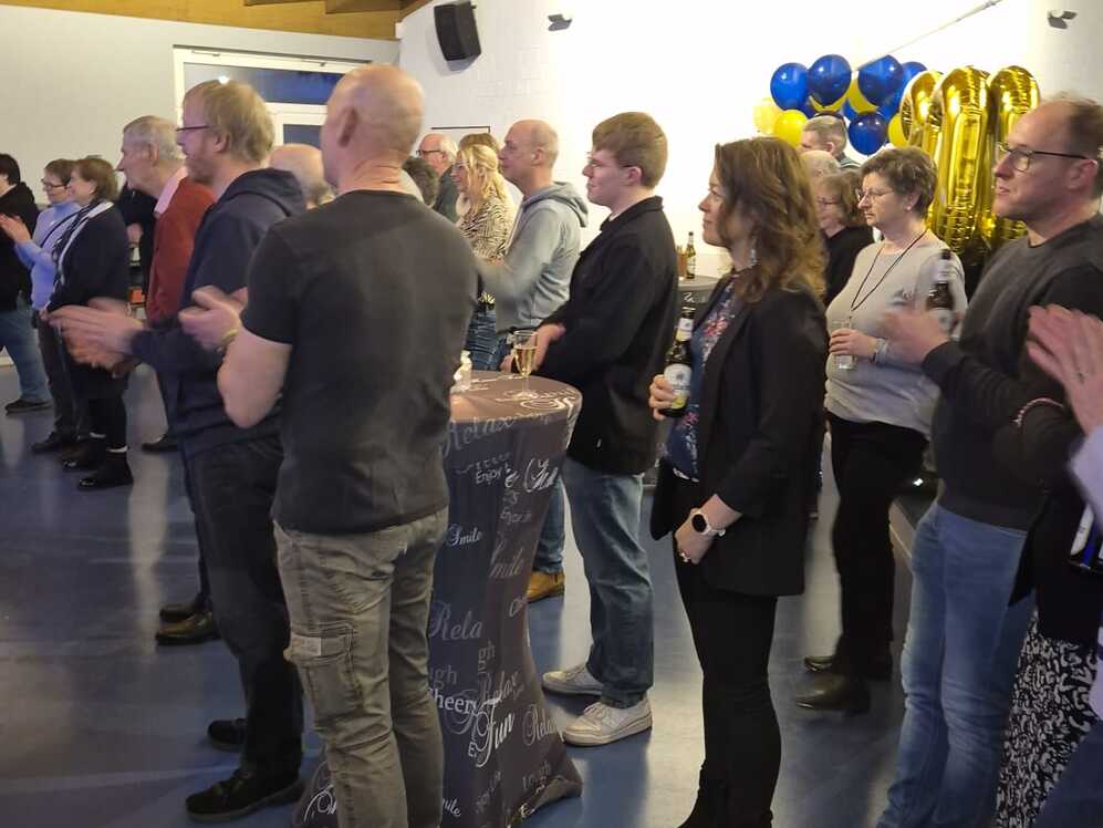 Viele Personen stehen in einem Raum mit blauem Fußboden und hören einem Vortrag zu.