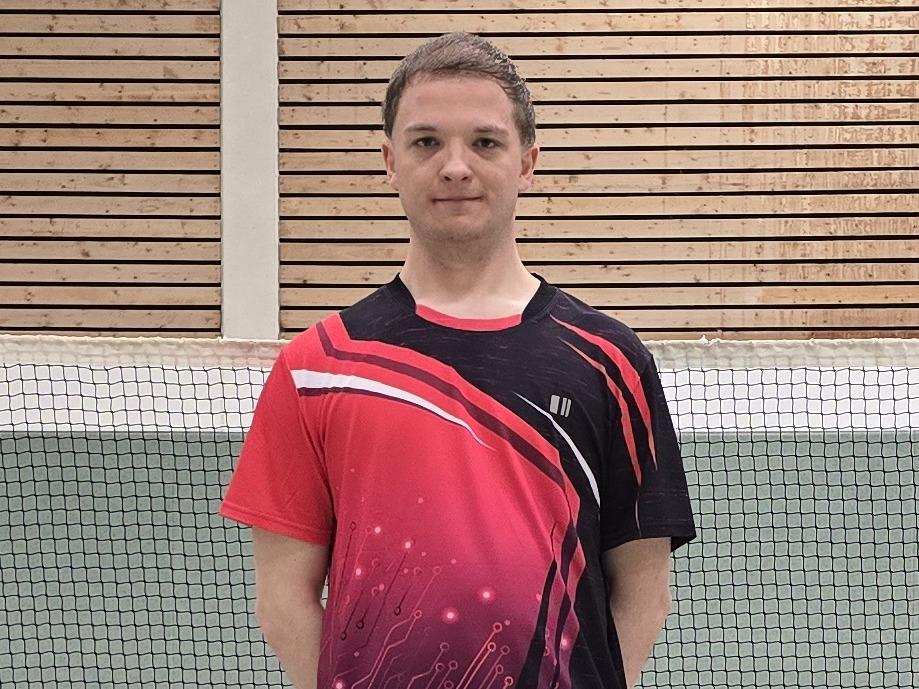 Ein Badmintonspieler in rotem Trikot steht auf einem Court, im Hintergrund ist das Netz und einige Federbälle verteilt.