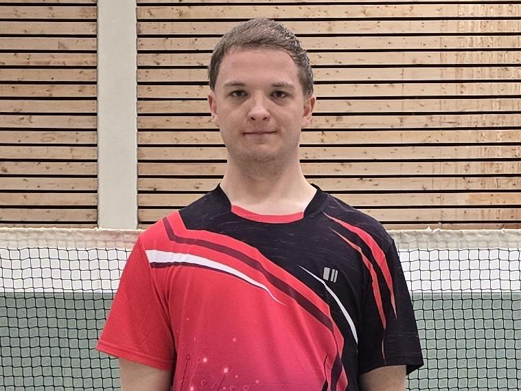 Ein Badmintonspieler in rotem Trikot steht auf einem Court, im Hintergrund ist das Netz und einige Federbälle verteilt.