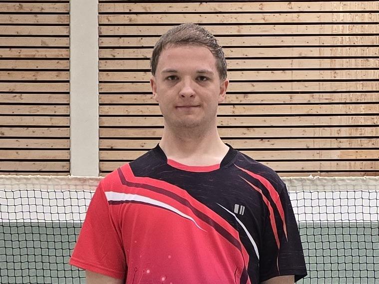 Ein Badmintonspieler in rotem Trikot steht auf einem Court, im Hintergrund ist das Netz und einige Federbälle verteilt.