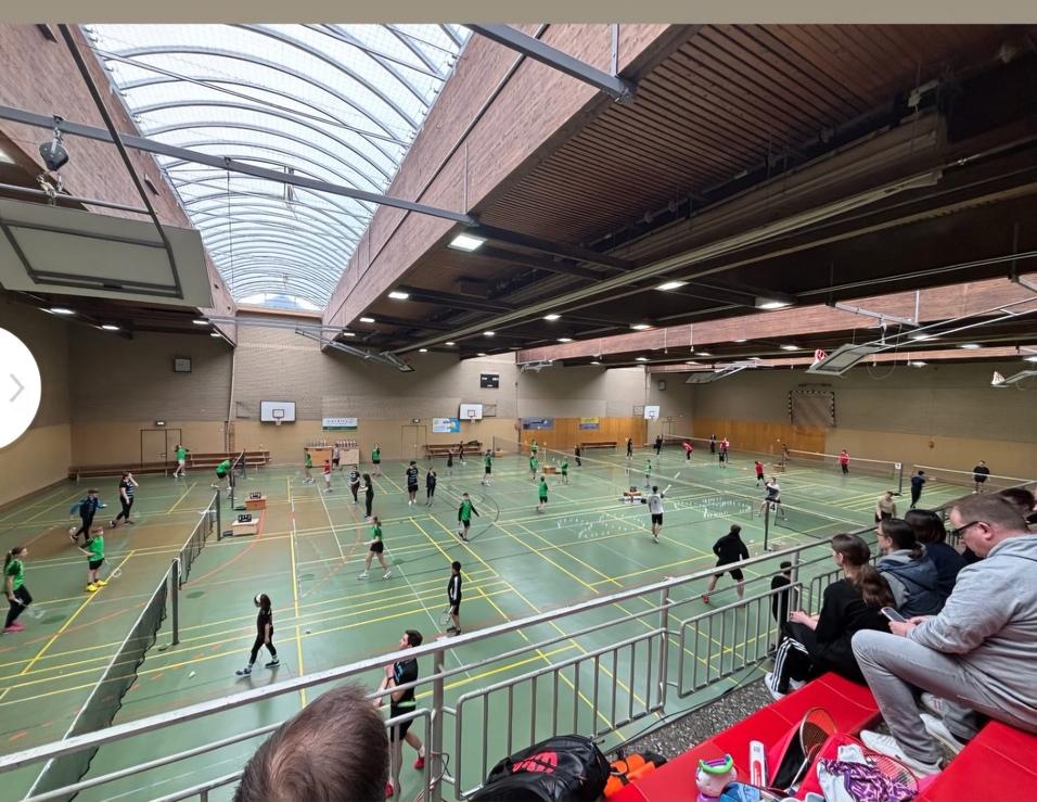 Große Sporthalle mit mehreren Badmintonspielern in grünen Trikots, Zuschauer sitzen auf der Tribüne.
