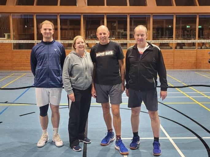 Vier Personen in Sportkleidung stehen in einer Halle auf einem Spielfeld mit Linien und einem Netz im Hintergrund.