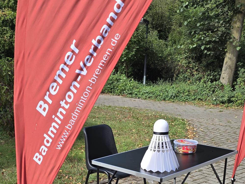 Ein Tisch mit einem übergroßen Badmintonball und einer Schale steht neben zwei Stühlen auf Pflastersteinen, links zwei rote Beachflags mit der Aufschrift 'Bremer Badminton-Verband' und einer Webadresse.