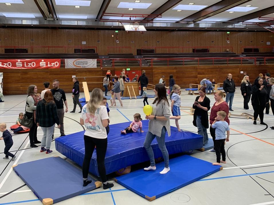Menschen und Kinder spielen in einer Sporthalle mit einer blauen Turnmatte und verschiedenen Spielgeräten.