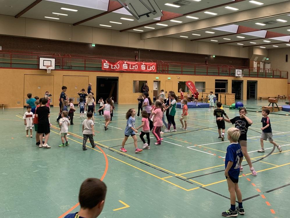 Kinder spielen und bewegen sich in einer großen Sporthalle mit Basketballkörben und einer Tribüne im Hintergrund.
