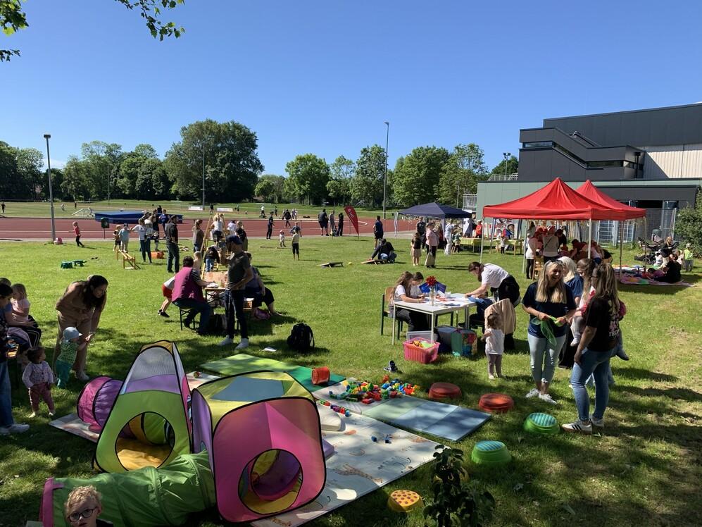 Mehrere Erwachsene und Kinder auf einer Wiese mit Spielzeug, Tischen und Pavillons bei einer Freiluftveranstaltung.