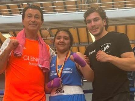 Drei Personen stehen in einem Boxring, die mittlere Person trägt Boxkleidung, Boxhandschuhe und eine Medaille, die beiden anderen tragen Sportkleidung.