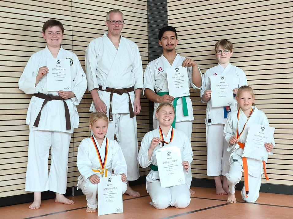 Gruppe von Karateka in weißen Kimonos hält Urkunden; einige tragen Medaillen, vor einer Holzpaneelwand.