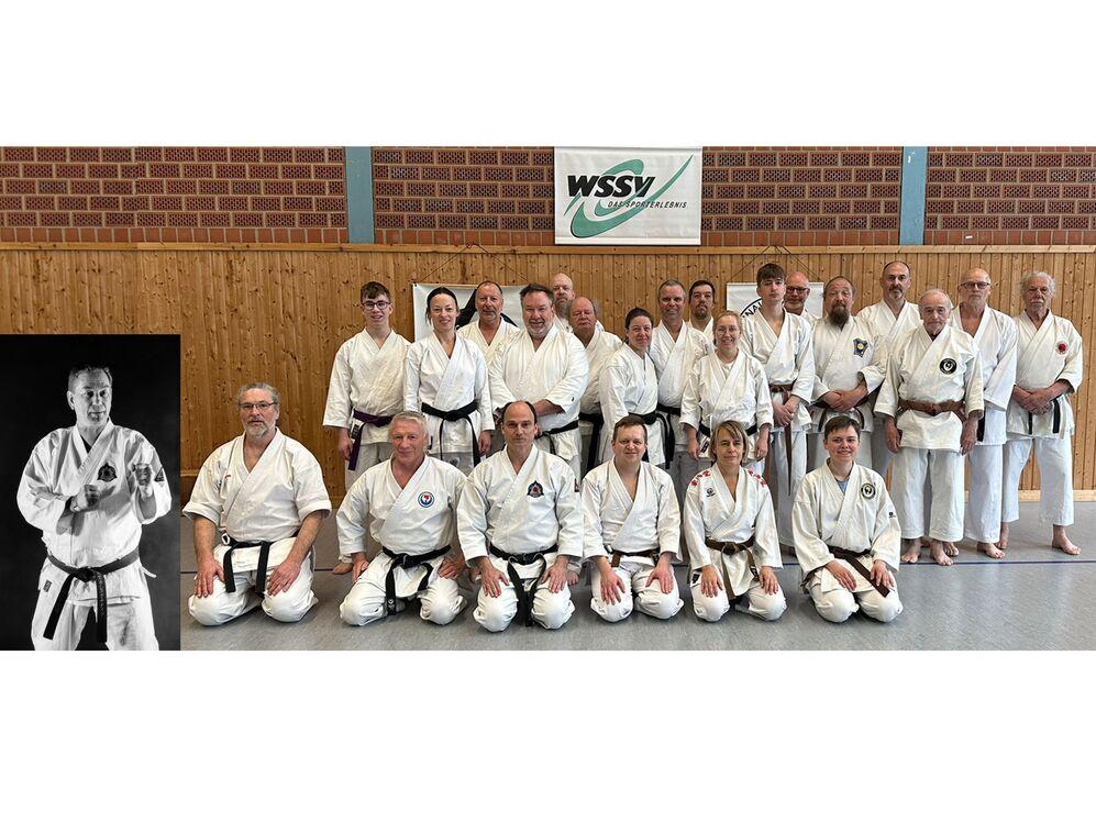 Gruppe von Erwachsenen in weißen Karateanzügen mit schwarzen Gürteln in einer Sporthalle vor einer Wand mit Holzverkleidung und einem Banner mit dem Schriftzug 'WSSV'.