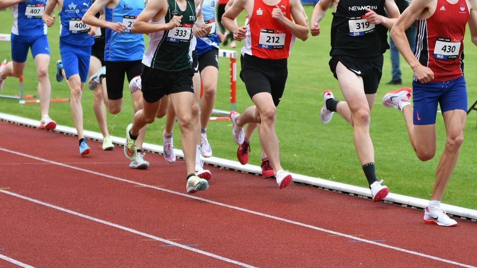 Sechs Läufer in Sportkleidung während eines Wettkampfs auf einer Laufbahn, im Hintergrund Zuschauer und Lautsprecher.