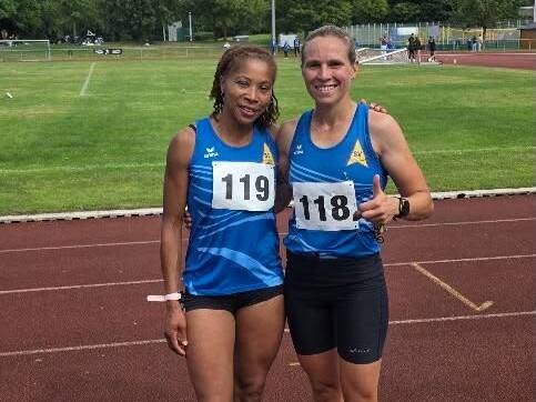 Zwei Sportlerinnen in blauen Trikots mit Startnummern stehen auf einer Laufbahn und lächeln in die Kamera.