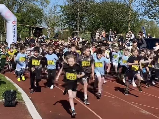 Gruppe von Kindern startet einen Lauf auf einer roten Laufbahn unter blauem Himmel. Zuschauer stehen im Hintergrund.