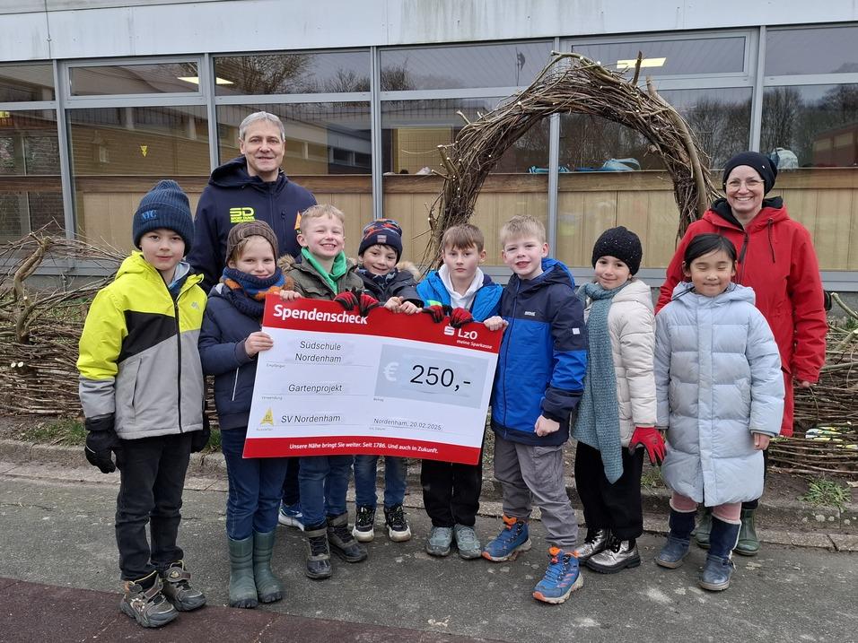 Gruppenbild von Kindern und Erwachsenen, die einen Spendenscheck für ein Gartenprojekt halten. Hintergrund: Schule.