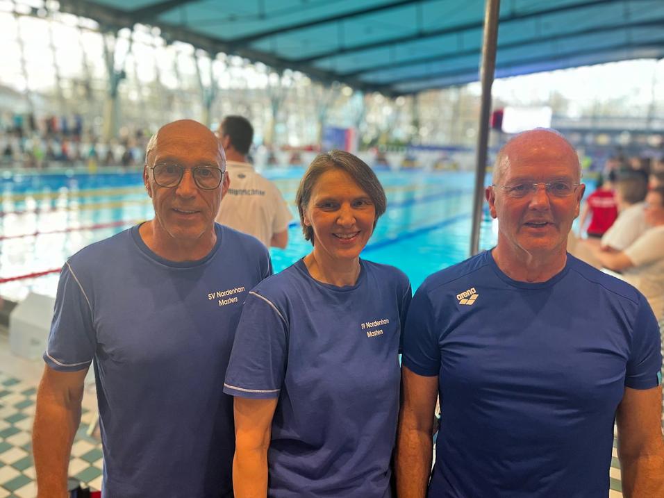 Drei Schwimmer in blauen T-Shirts stehen vor einem Schwimmbecken in einem Wettkampfumfeld.