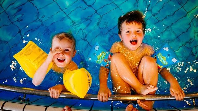 Zwei fröhliche Kinder mit Schwimmhilfen im Wasser, lächeln und halten sich am Rand des Schwimmbeckens fest.
