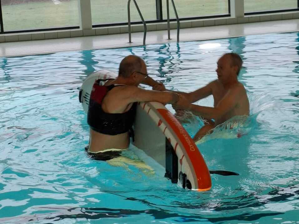 Zwei Personen im Schwimmbecken behelfen sich beim Umgang mit einem Surfbrett. Sichtbare Fenster und ein gelbes Fahrzeug im Hintergrund.