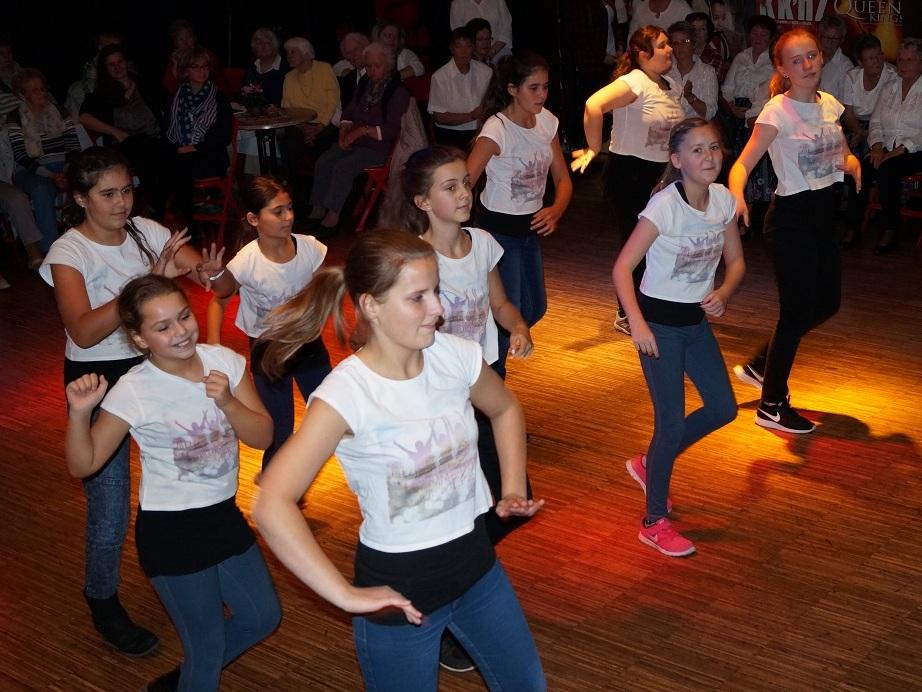 Tanzgruppe aus Mädchen in weißen Shirts und dunklen Hosen führt eine Choreografie vor Publikum auf einer Theaterbühne auf.