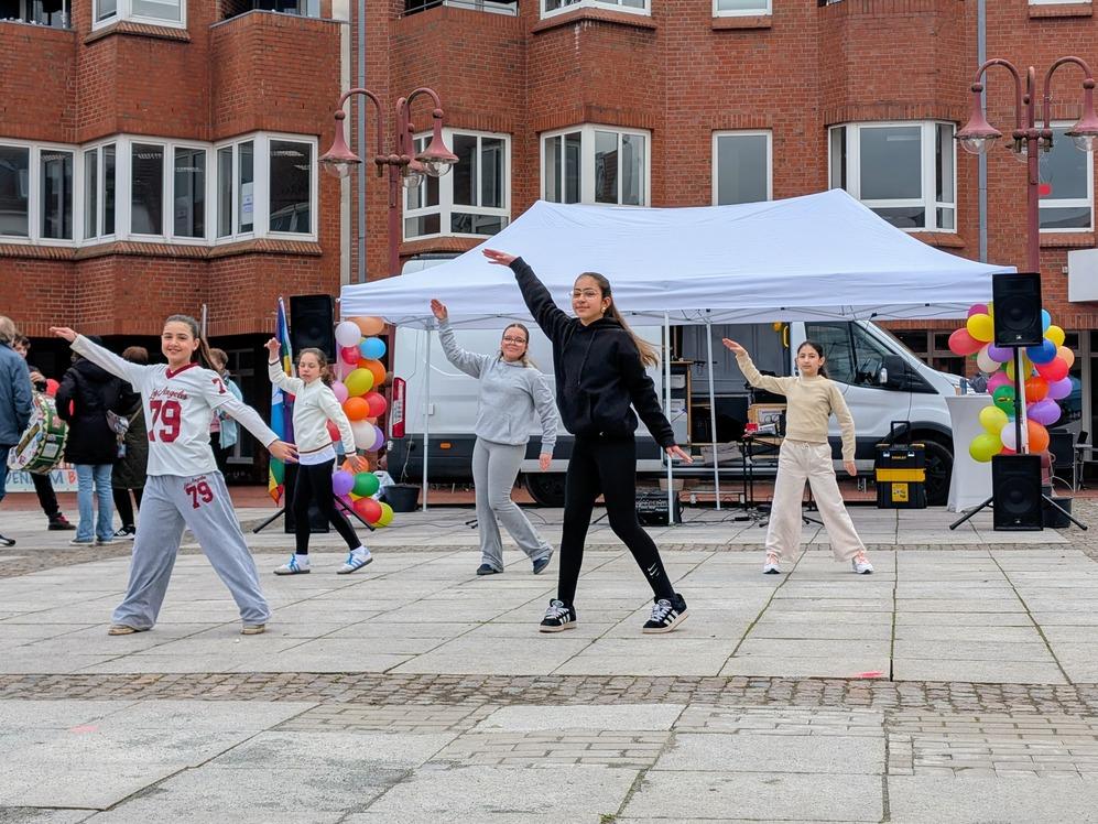 Fünf junge Frauen tanzen auf einem gepflasterten Platz vor einem weißen Zelt und einem roten Backsteingebäude.