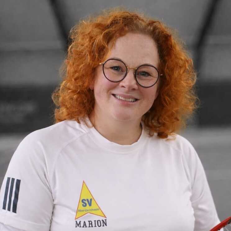 Porträt einer lächelnden Frau mit rötlichen Haaren, Brille und weißem Oberteil, vor einem unscharfen Hintergrund.