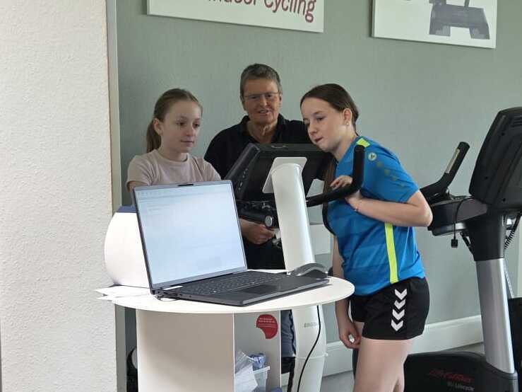 Drei Personen in einem Fitnessraum vor einem Laufband, zwei sitzen an einem Tisch mit Laptop, eine steht und zeigt auf den Bildschirm.