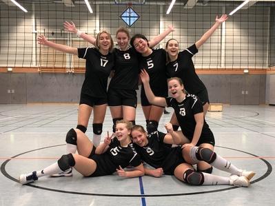 Gruppensfoto von sechs Volleyballspielerinnen in schwarzen Trikots, fröhlich posierend in einer Sporthalle.
