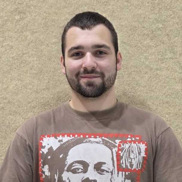 Junger Mann mit Kurzhaarschnitt, trägt ein T-Shirt mit Aufdruck, hält einen Ball in beiden Händen.