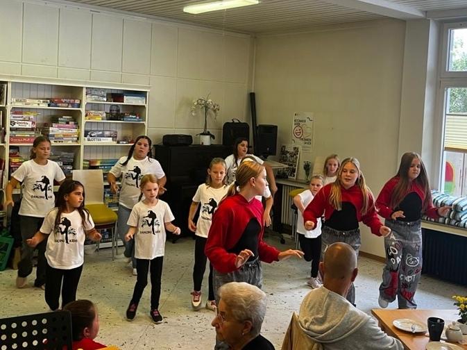Kinder tanzen vor einer Gruppe älterer Menschen in einem Gemeinschaftsraum mit Bücherregal und großen Fenstern.